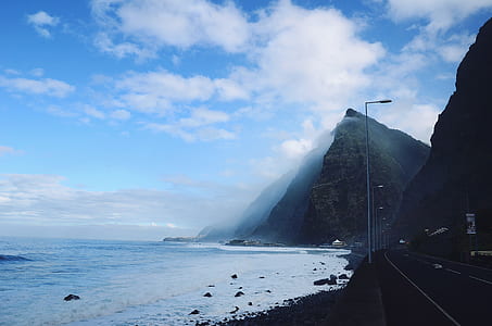 gray roadway near ocean and mountain under blue and cloudy sky thumbnail