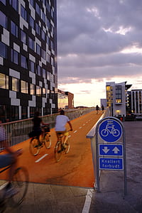 two person riding bicycle near grey concrete building under cloudy sky thumbnail