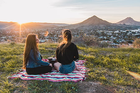 two women sitting on multicolored mat near mountain during daytime thumbnail