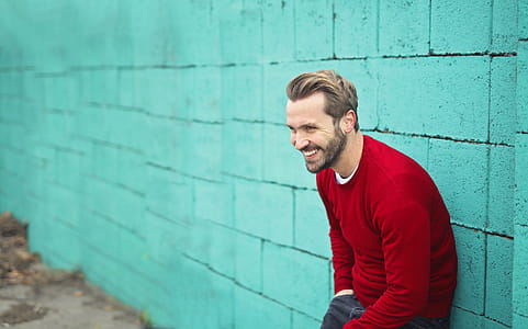 Young Adult Man wearing a red sweater leaning on a turquoise brick wall thumbnail
