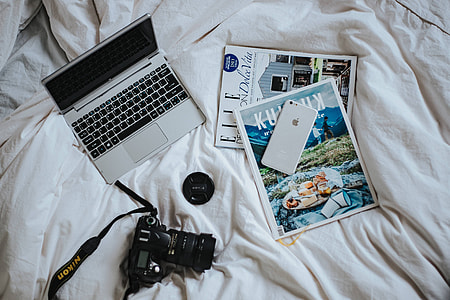 Silver laptop, a camera, magazines and other items on white bed sheets thumbnail