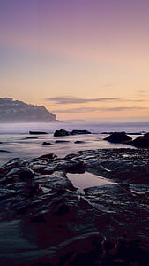 black and gray big rocks beside body of water near mountain under gray sky at golden hour thumbnail