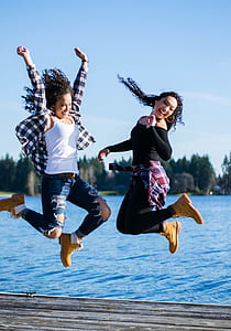 two women with curly hairs jumping on gray wooden platform while smiling near body of water during daytime thumbnail