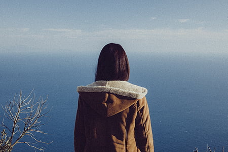 woman wearing brown leather hooded jacket overlooking blue calm body of water during daytime thumbnail