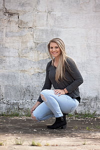 woman wearing grey long-sleeved shirt and distressed blue denim skinny jeans and black buckled boots kneeling near grey concrete wall thumbnail