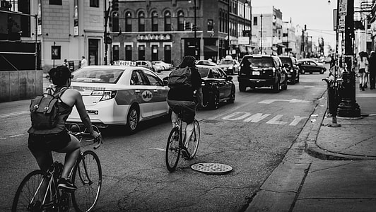 greyscale photo of people in bike riding along cars on road with buildings thumbnail