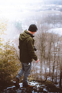 man in black knit cap, green hooded jacket, blue denim jeans, and pair of brown boots standing on cliff during daytime thumbnail