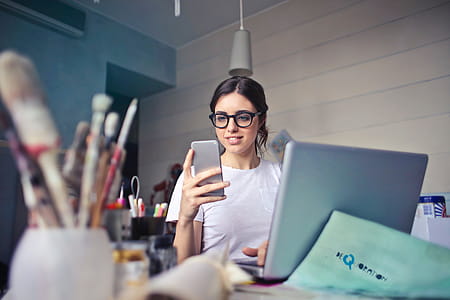 A young woman with glasses on her laptop looks at her mobile phone thumbnail
