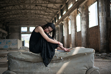woman sitting on sofa wearing black sleeveless maxi dress holding arms inside abandoned concrete building thumbnail