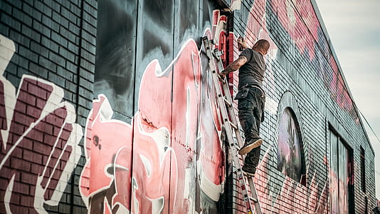 Man in Gray Shirt Standing on Gray Steel Ladder Painting Black White and Red Graffiti on Concrete Wall Outdoors thumbnail