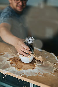 person holding clear glass jar filled with ice on white ceramic mug with spilled coffee on brown wooden table inside room thumbnail