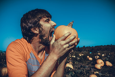 man wearing orange crew-neck shirt eating orange pumpkin during daytime photo thumbnail