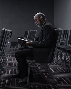 man wearing suit jacket sitting on metal chair while reading magazine thumbnail