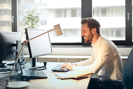Young Man In White Shirt Sitting on Black Rolling Chair While Facing Computer Set and Smiling thumbnail