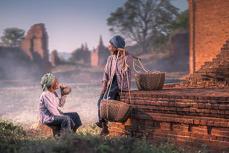 two women talking while sitting beside brown brick building during daytime thumbnail