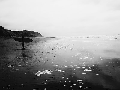 grayscale photography of person holding surfboard standing on shore thumbnail