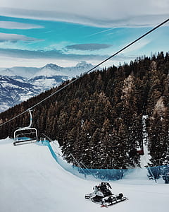 snow vehicle near trees below ski lift in distant of snow capped mountains thumbnail