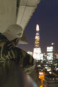 man wearing white curved brim cap while staring at skyscrapers during nighttime thumbnail