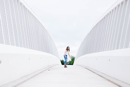 woman in white top and blue destressed jeans riding longboard beside white walls during daytime thumbnail