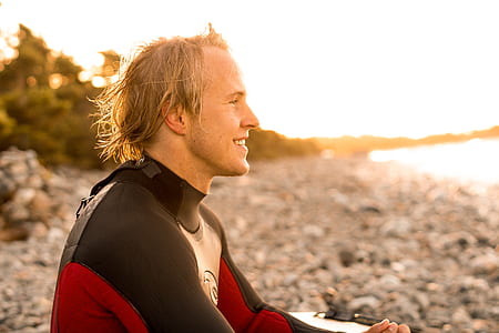 smiling man in red and black wetsuit sitting on seashore at daytime thumbnail
