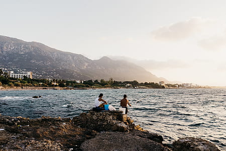 man sitting on stone near man standing on stone while fishing on body of water thumbnail