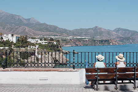 two persons sitting on brown bench facing buildings on hill beside body of water thumbnail