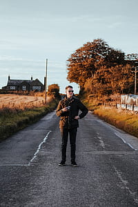 man wearing quilted brown bubble jacket holding camera standing on the road photography thumbnail