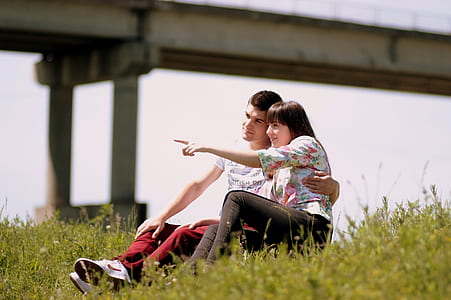 couple sitting on grass field while girl pointing something during golden hour thumbnail