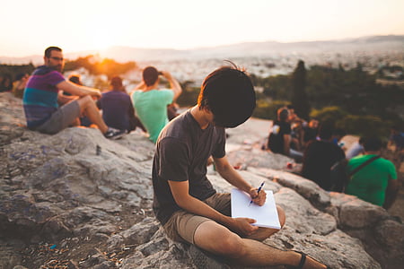 person holding pen writing on paper while sitting on rock formation take during daytime thumbnail