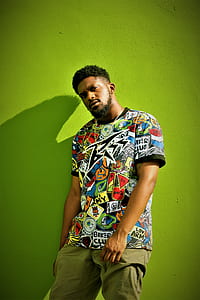 man leaning on green wall wearing multicolored crew-neck t-shirt and brown bottoms thumbnail