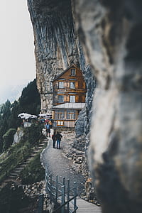 brown wooden house at the edge of hill surrounded with rocks and people during daytime thumbnail