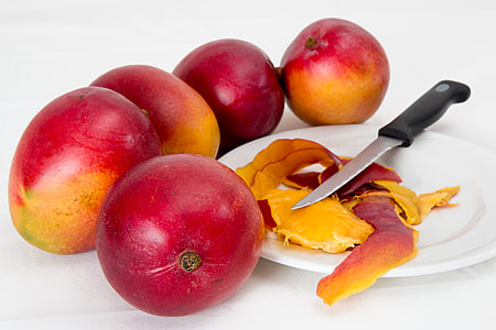 five red mangoes beside white plate with peeled mango and knife thumbnail