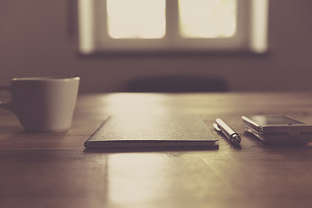 selective focus photo of book and pen on wooden table near mug and smartphone thumbnail