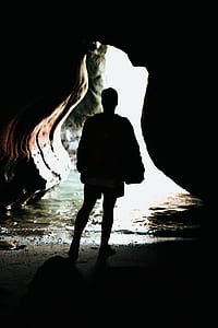 person wearing jacket standing in front of entrance of a cave during daytime thumbnail