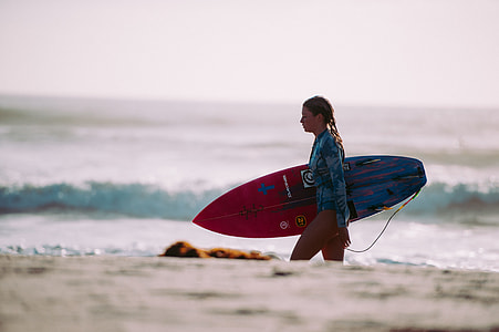 woman carrying surfboard walking on seashore during daytime thumbnail