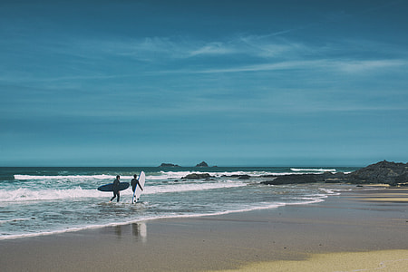 Two surfers walk along the beach on a sunny day on the coast of Cornwall in England thumbnail