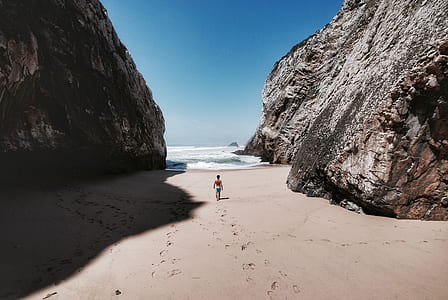 man walking toward sea on brown sand between rocks at daytime thumbnail