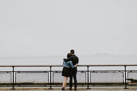 man and woman hugging beside black concrete fence in front of body of water during daytime thumbnail