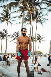 man wearing red shorts standing on concrete fence near coconut tree on seashore thumbnail