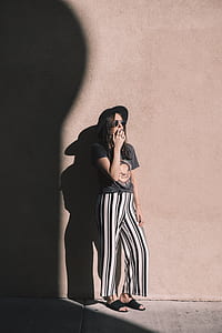 woman wearing black and beige t-shirt, black hat, and white and black striped pants holding smoke and leaning behind beige wall thumbnail