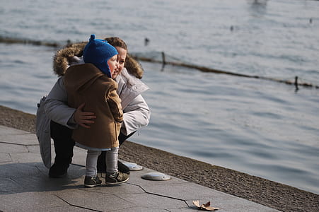 boy standing beside crouching person in front of body of water during daytime thumbnail