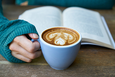 person holding blue ceramic cup with coffee inside and book behind thumbnail