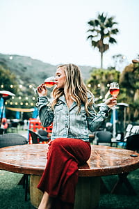 woman holding two drinking glass of liquors sitting on brown wooden table at daytime selective focus photography thumbnail
