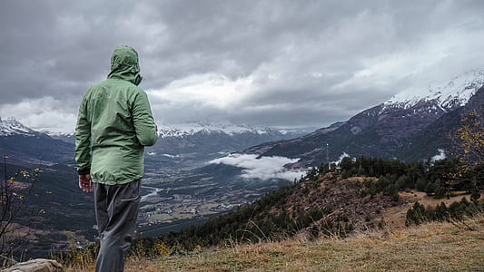 person wearing green jacket and gray pants standing in front mountain thumbnail