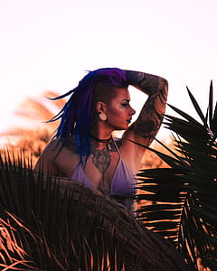 woman in purple bikini top standing behind trunk and leaves raising her left hand thumbnail
