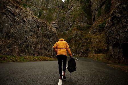 photo of person wearing yellow hoodie and black pants holding backpack walking on roadway between mountain thumbnail
