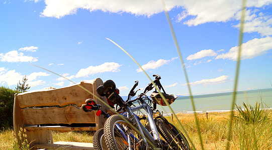 two grey hard-tail mountain bike with brown wooden bench in front of a calm sea under the blue sky and white clouds during daytime thumbnail