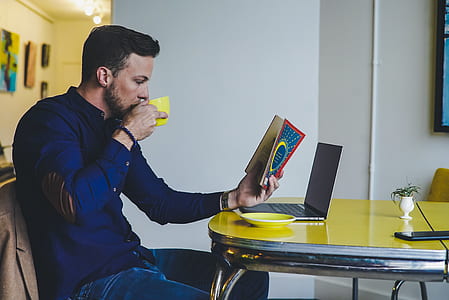 man in blue dress shirt holding book and yellow teacup in front of laptop computer thumbnail