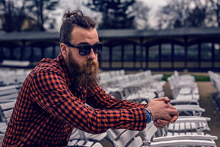 Man Wearing Black and Red Checkered Long Sleeve Shirt Wearing Black Wayfarer Sunglasses Sitting on White Wooden Chair thumbnail