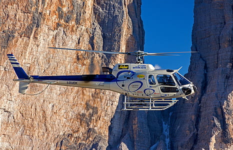 white and blue helicopter flies near brown rock formation thumbnail